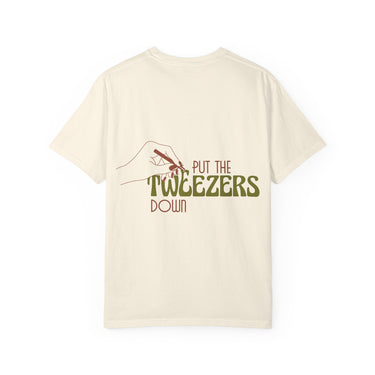 Cream t-shirt featuring a back graphic of a reddish-brown outlined hand holding tweezers and the phrase PUT THE TWEEZERS DOWN with TWEEZERS in an olive green groovy font
