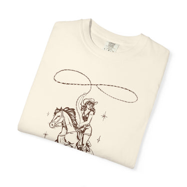 Cream Comfort Colors t-shirt displaying a vintage-style pin-up cowgirl in a cowboy hat and sunglasses, wearing a star-patterned top with the number 29, riding a horse and swinging a brown outline lasso, accented with subtle starbursts