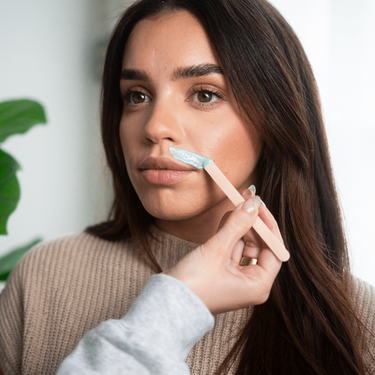 Dark-haired woman in a beige knitted sweater getting her upper lip waxed with light blue wax on a wooden stick in a bright salon setting with green foliage