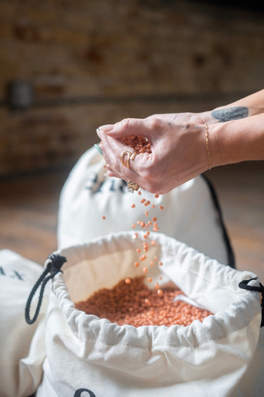 Close-up of hands with light green nail polish and gold rings pouring reddish-brown seeds into a white drawstring bag indoors