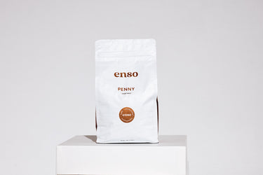 White Enso Penny Hard Wax product pouch displayed on a white plinth in a minimalist studio setting showing its brand logo, 'PENNY HARD WAX' text, and a circular brown 'vegan antimicrobial hypoallergenic' label