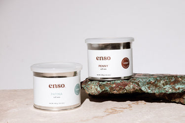 Product photo of two enso soft wax containers, 'Penny' featuring brown branding on a rustic copper block with vibrant teal and rust patina, and 'Patina' with teal branding on a cream marble surface