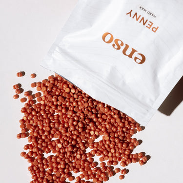 A white pouch of ENSO PENNY HARD WAX spilling numerous shimmering coral-colored wax beads onto a bright white surface