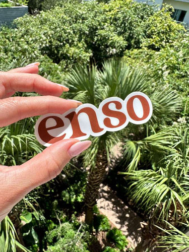 Photograph of a hand with manicured nails holding a white sticker featuring rust-brown Enso text against a vibrant background of lush green palm trees and dense sunlit foliage