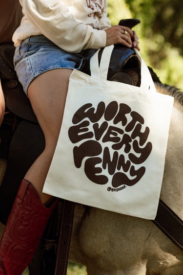 Close-up of a person riding a light brown horse outdoors, holding a cream canvas tote bag featuring 'WORTH EVERY PENNY @ensouwax' in brown bubble letters, wearing denim shorts and vibrant red cowboy boots