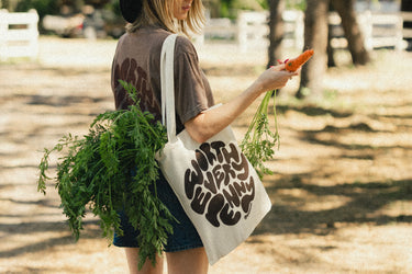 Photograph of a person holding a vibrant orange carrot with green tops, featuring red fingernails and a gold ring, while carrying a beige canvas tote bag with dark brown 'Grow Your Own' lettering, in a rural setting with blurred white fences and trees
