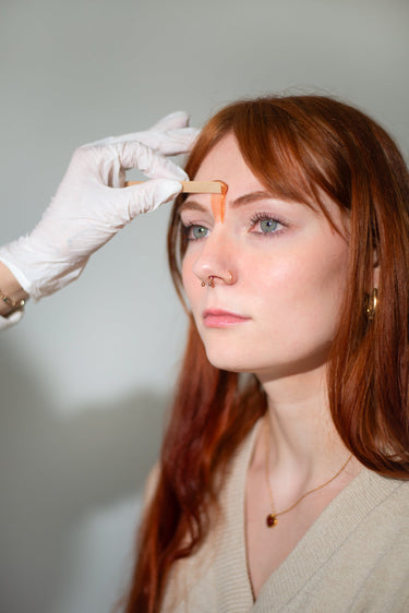 Portrait of a redhead woman with striking blue-green eyes and gold nose piercings getting an orange wax applied to her forehead by a gloved hand with a wooden stick