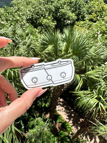 Photograph of a hand with light pink manicured nails holding a black and white outline sticker of a dual slow cooker against a vibrant outdoor backdrop of green tropical palm trees and dense foliage