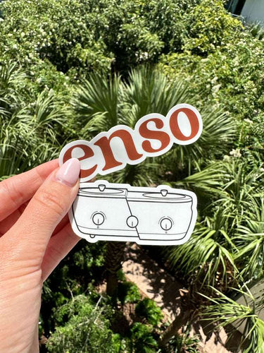 Photograph depicting two white stickers, one with reddish-brown 'enso' text and the other a line drawing of two connected slow cookers, held by a hand with light pink nails against a vibrant green tropical foliage background including palm fronds