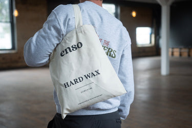 Photograph of a person from behind wearing a gray sweatshirt with visible green text, carrying a cream canvas tote bag labeled 'enso HARD WAX 10 LBS | 4.5 KG' in an industrial loft with large windows and a wooden floor