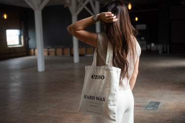 Photograph of a woman from behind, with long brown hair, wearing a beige knit top and white pants, holding a cream canvas tote bag with "enso HARD WAX" text, standing in an expansive industrial space with white columns and warm glowing lightbulbs