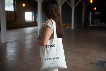 A woman with brown hair and a beige knitted top holds a white enso Hard Wax tote bag inside a spacious industrial loft featuring warm ceiling lights and wooden floors